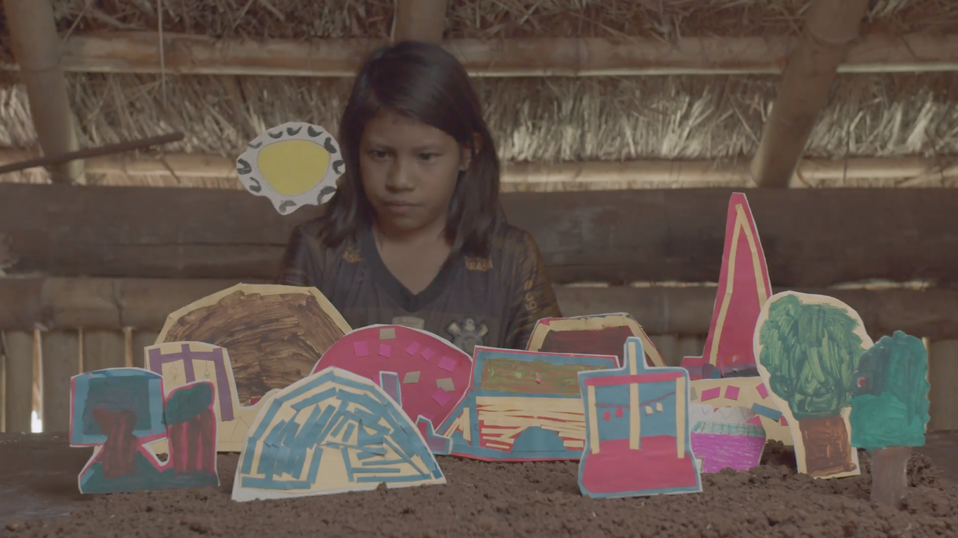 Menina indígena observa maquete colorida de papel em uma mesa de terra, com figuras representando casas, árvores e o sol ao fundo, em uma estrutura de madeira com teto de palha.